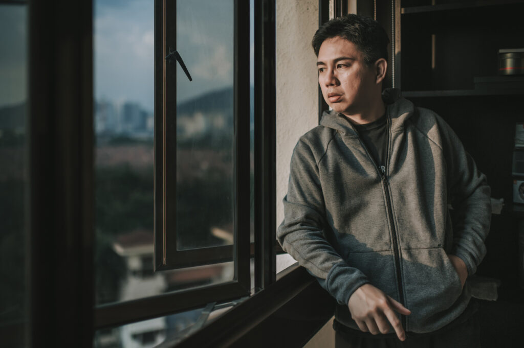 an asian chinese mid adult man looking outside of window from his home during sunset with serious facial expression with hooded shirt malaysian-graduates-job-mismatch-and-unemployment-challenges